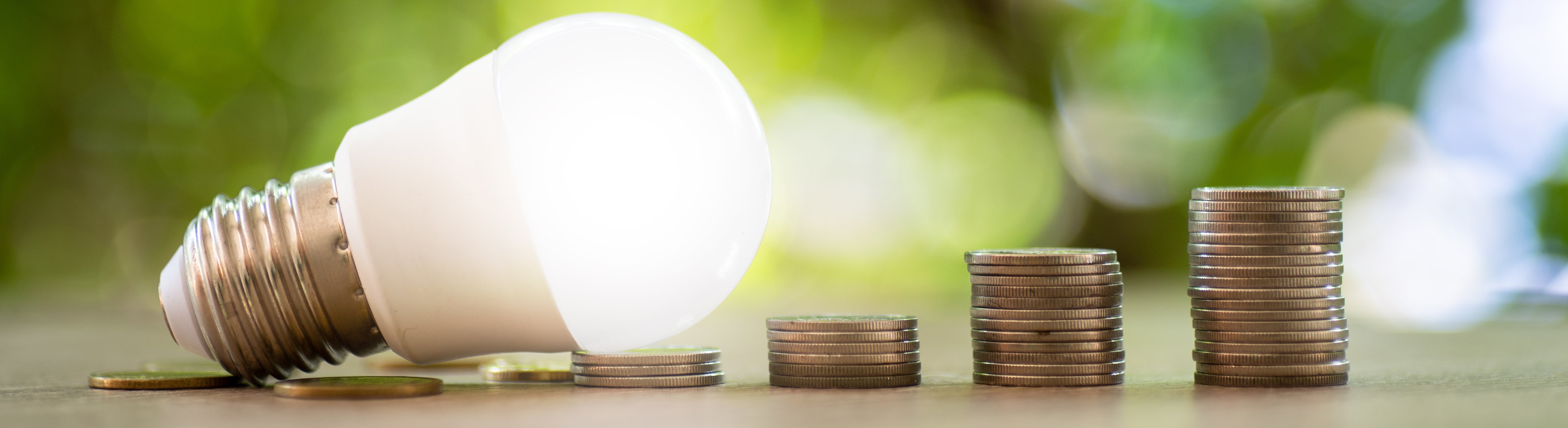 A glowing LED light bulb leans over piles of coins