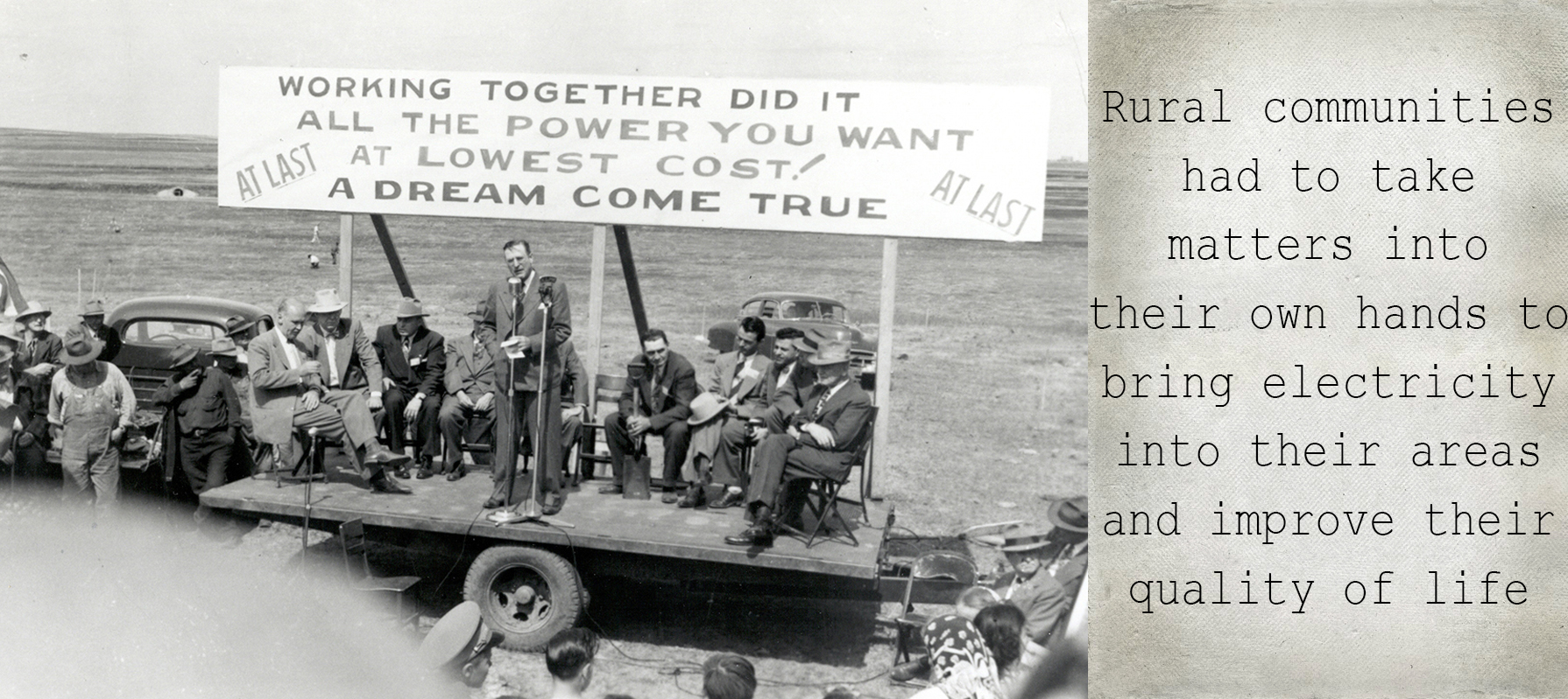 Rural communities had to take matters into their own hands to bring electricity into their areas and improve their quality of life; image shows group of men in suits sitting on platform, with a man standing in front of them at a microphone and a sign behind them, reading "Working together did it, all the power you want at lowest cost! At last, a dream come true."