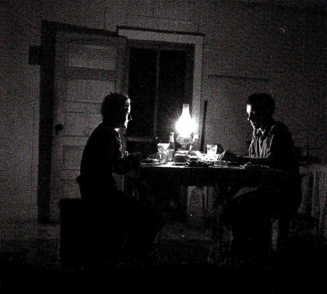 Newlywed couple sitting in a darkened room, lit only by a dim oil lamp. Photo Source: "The Next Greatest Thing," published by NRECA. Original Photo by Esther Bubley, entitled "Newlyweds" from the Standard Oil of New Jersey college
