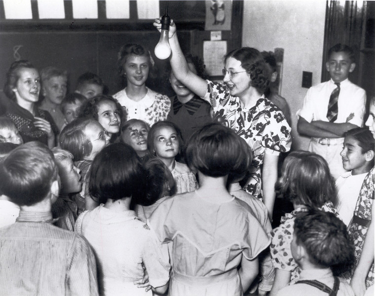 A teacher holds onto an electric lightbulb hanging from the ceiling in her schoolhouse, as interested children gather around.  Source: The Next Greatest Thing, published by NRECA