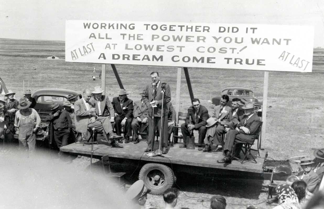A presentation by REA officials lets new member-owners know that their combined efforts were responsible for their rural areas being electrified.  The sign on the platform reads: “WORKING TOGETHER DID IT! ALL THE POWER YOU WANT AT LOWEST COST! A DREAM COME TRUE! AT LAST! AT LAST!”  Source: The Next Greatest Thing, published by NRECA