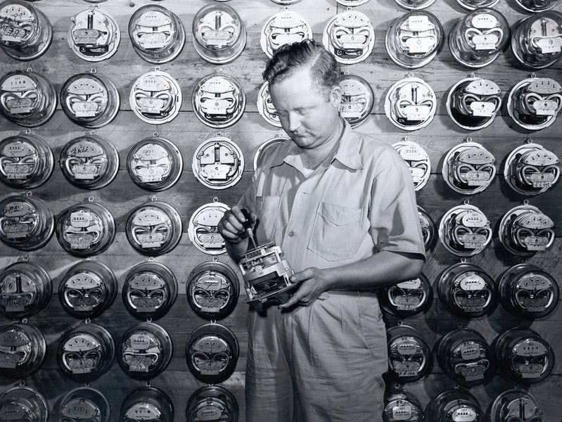 Black-and-white photo of a meter technician tinkering with an electric meter. Source: “The Next Greatest Thing,” published by NRECA