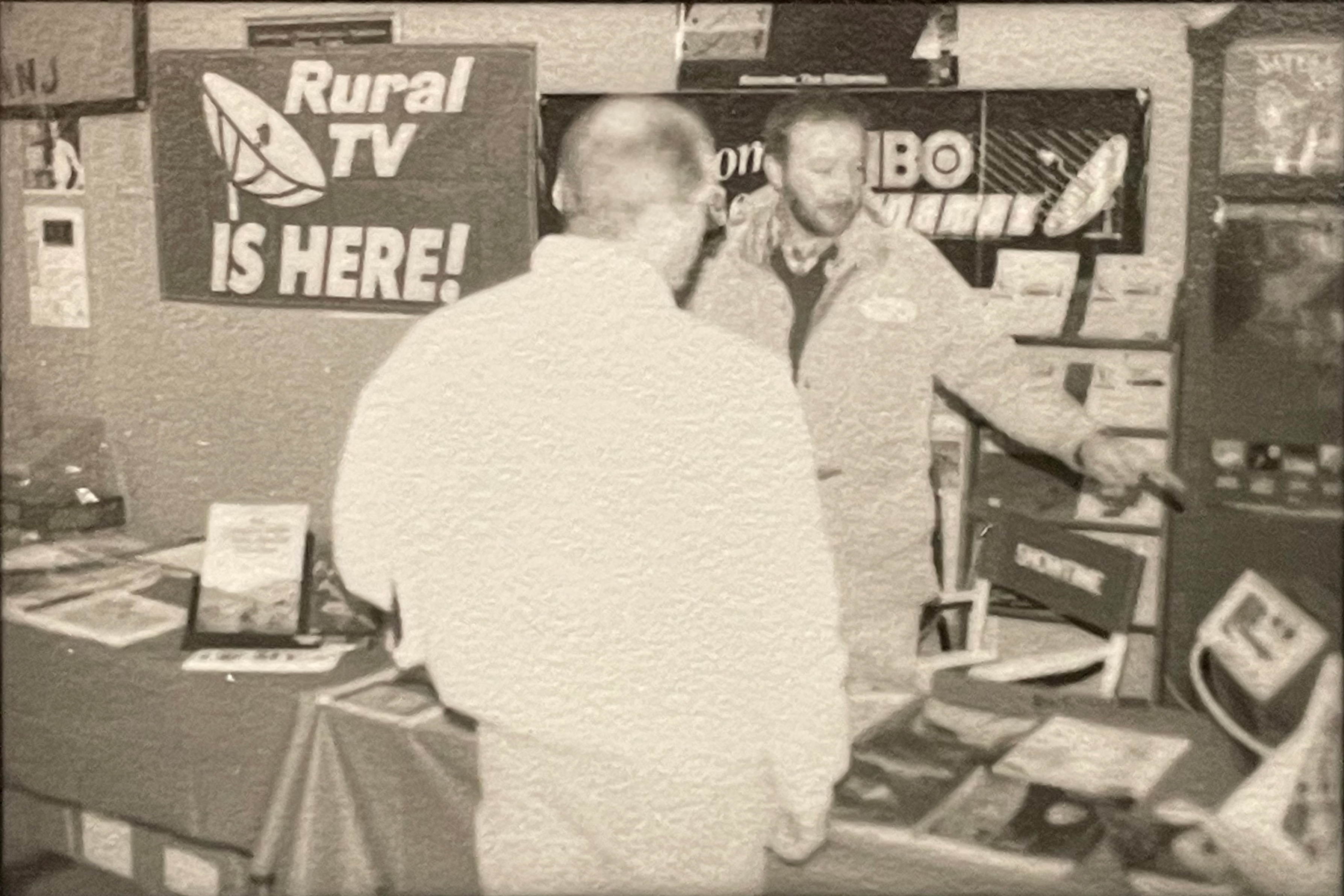 Pictured: Two men speaking at a public event discussing the benefits of satellite TV for rural communities. A prominent sign in the background reads: “Rural TV IS HERE!”