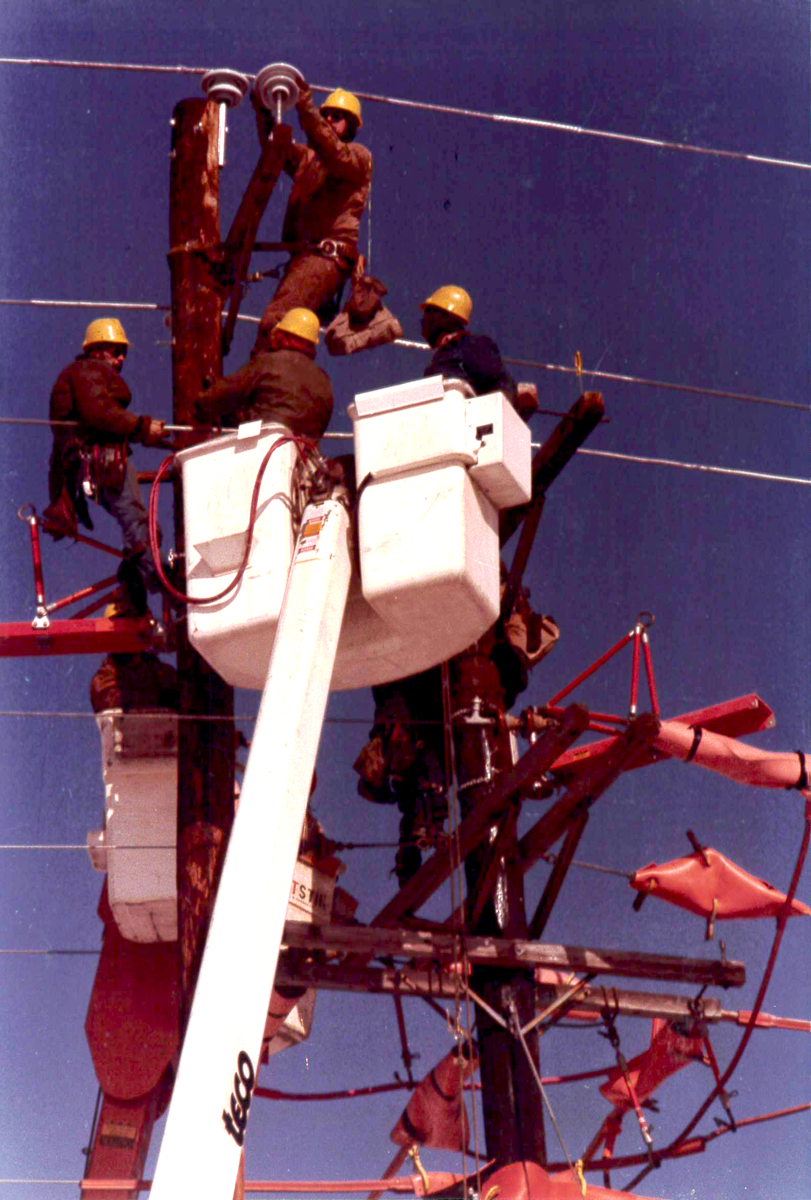 A vintage photo depicting a group of linemen from Sussex Rural Electric Cooperative performing work on a pole, changing out reclosers. Two of the linemen had climbed the pole to perform this work, with support from the other two working out of a bucket. They are making use of line covers to protect themselves from shock.